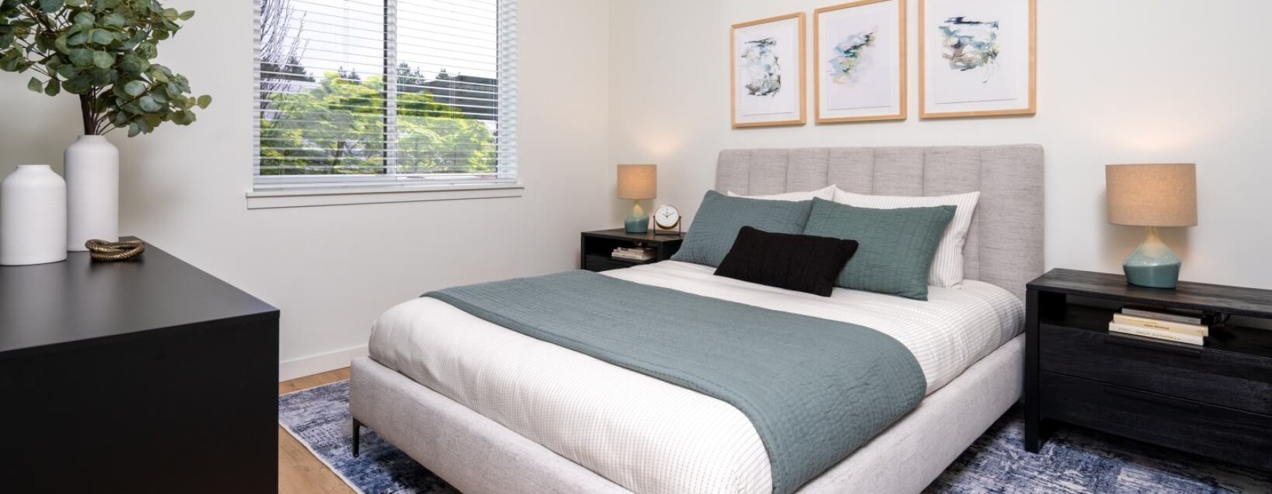 Bright bedroom with a modern upholstered bed, blue-gray bedding, framed wall art, and a window with natural light.