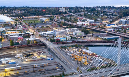  Aerial dusk view of a mid-sized city featuring a cable-stayed bridge in the foreground, an active railway yard, a large domed arena, highway interchanges, and a mix of commercial and residential buildings stretching into the distance.