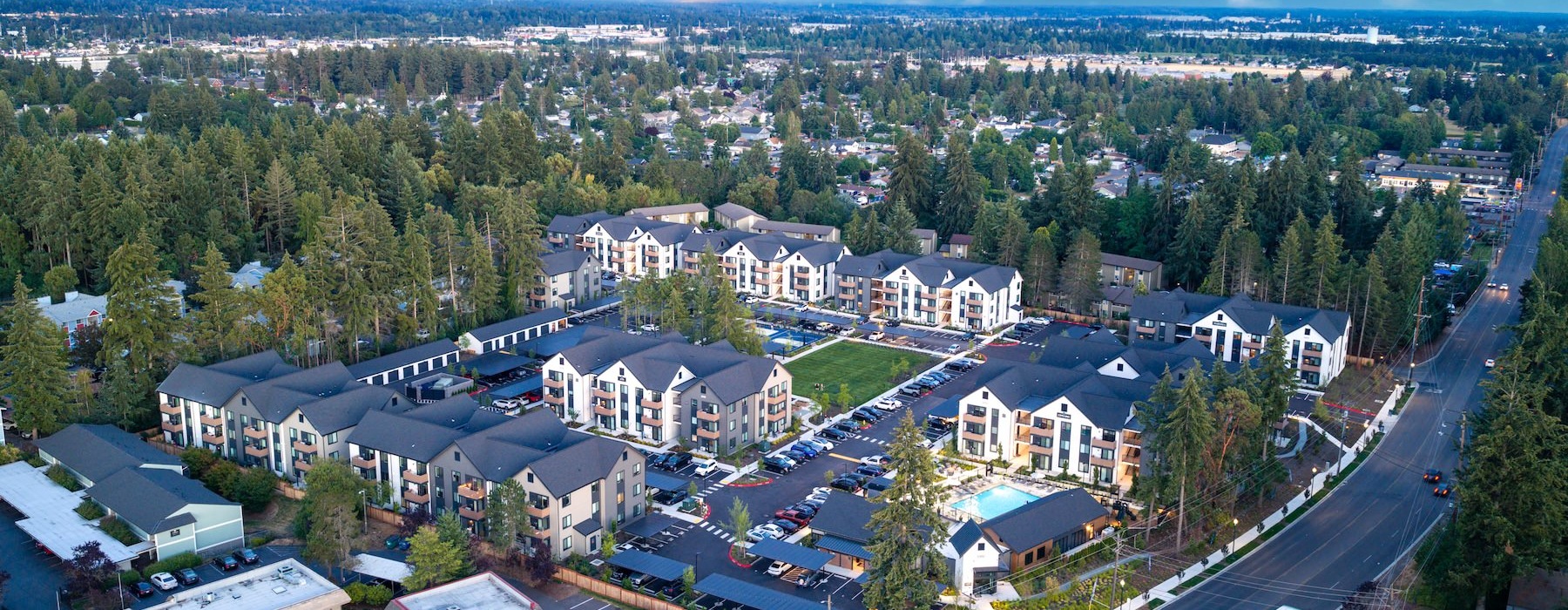 Aerial view of The Rigney Apartments complex surrounded by trees, with a pool and parking areas, at dusk.