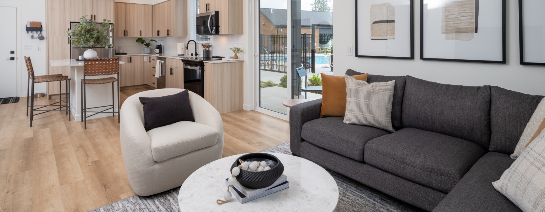 A modern open-concept apartment living room with a dark gray sofa, cream accent chair, and marble coffee table, flowing into a light wood kitchen with a sliding glass door overlooking a pool area.