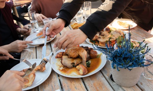 A group of people dining outdoors at a rustic wooden table, sharing plates of roast chicken with gravy, mashed potatoes, and vegetables, with a small blue flower arrangement as a centerpiece.
