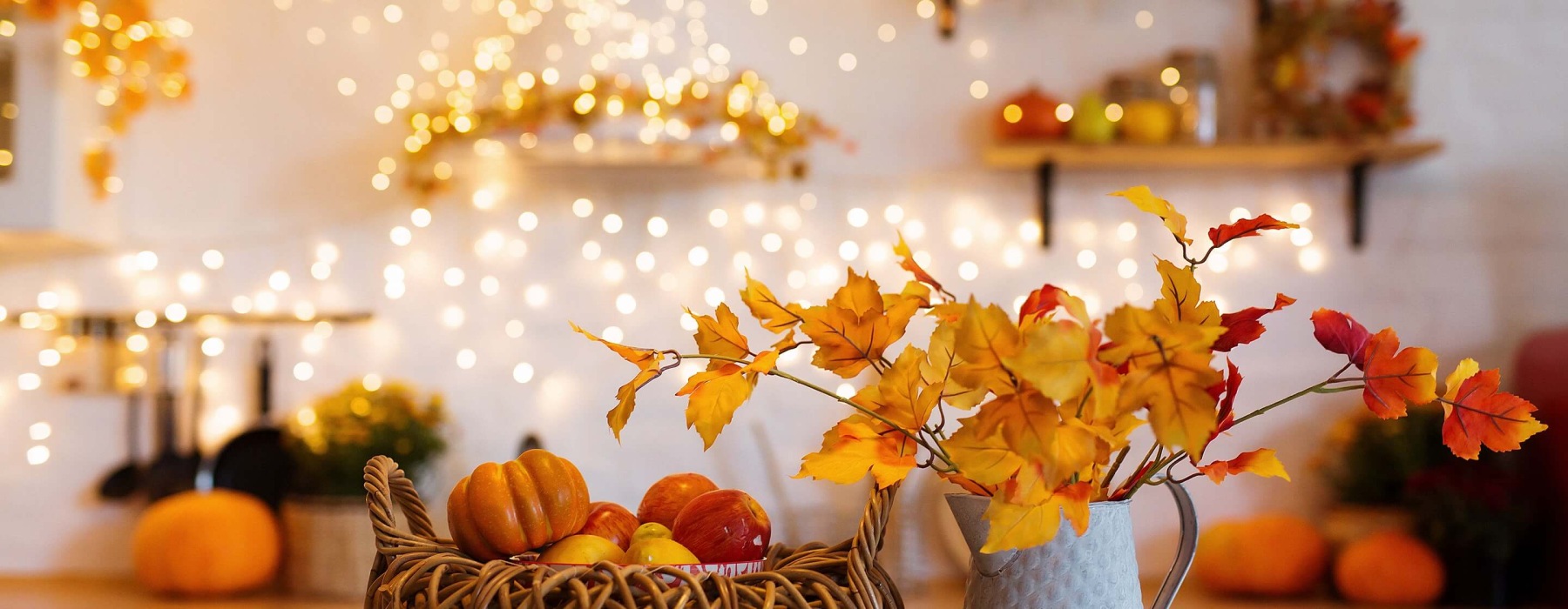 A kitchen counter decorated for fall with a woven basket of mini pumpkins and apples, a metal pitcher holding autumn leaf branches, and warm string lights glowing in the blurred background.