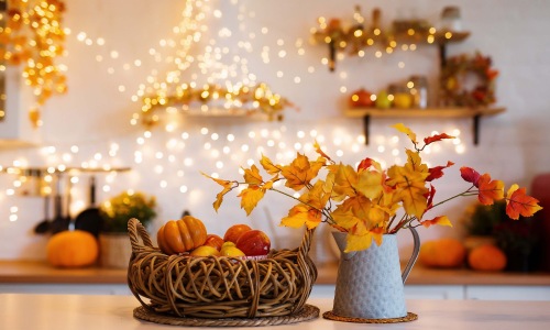 A kitchen counter decorated for fall with a woven basket of mini pumpkins and apples, a metal pitcher holding autumn leaf branches, and warm string lights glowing in the blurred background.