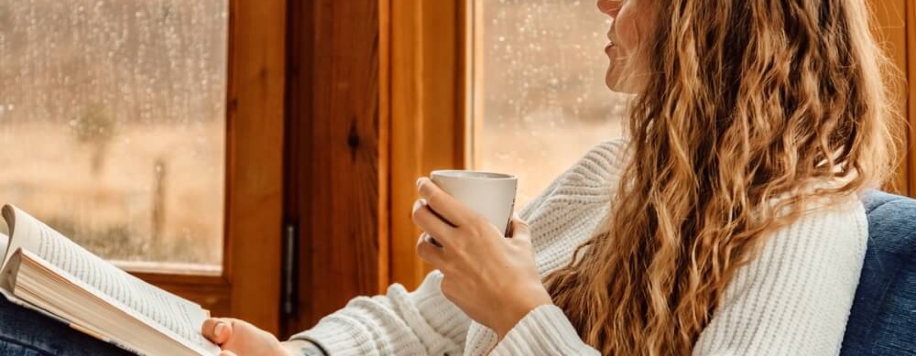 A woman with curly hair relaxes by a rain-streaked window, holding a mug and an open book.
