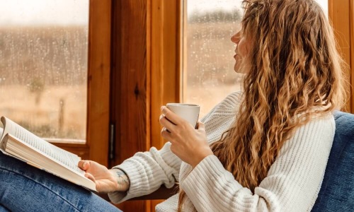 A woman with curly hair relaxes by a rain-streaked window, holding a mug and an open book.